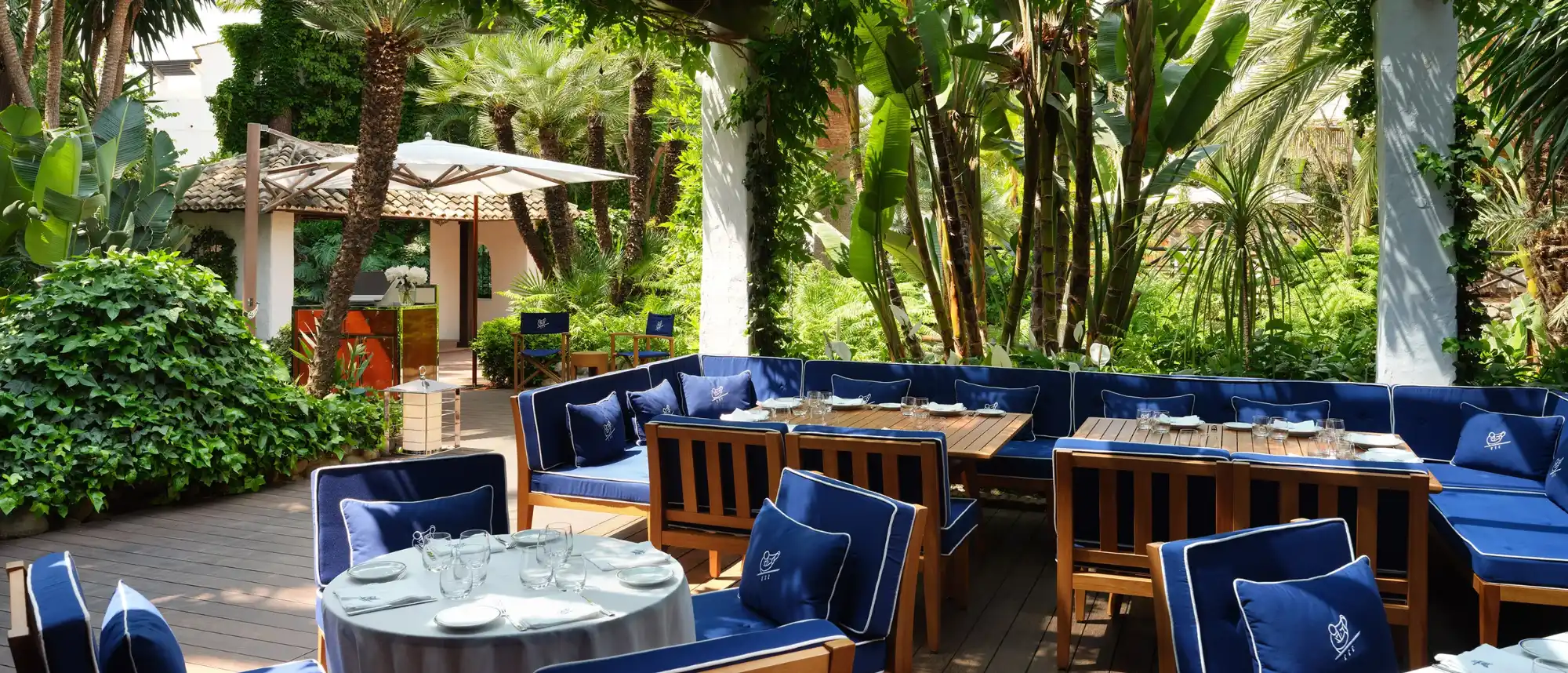 The elegant, shaded garden terrace of Cipriani Marbella at Puente Romano Beach Resort, featuring signature deep blue cushions with the white Cipriani logo, wooden tables, and director's chairs surrounded by luxury pool views